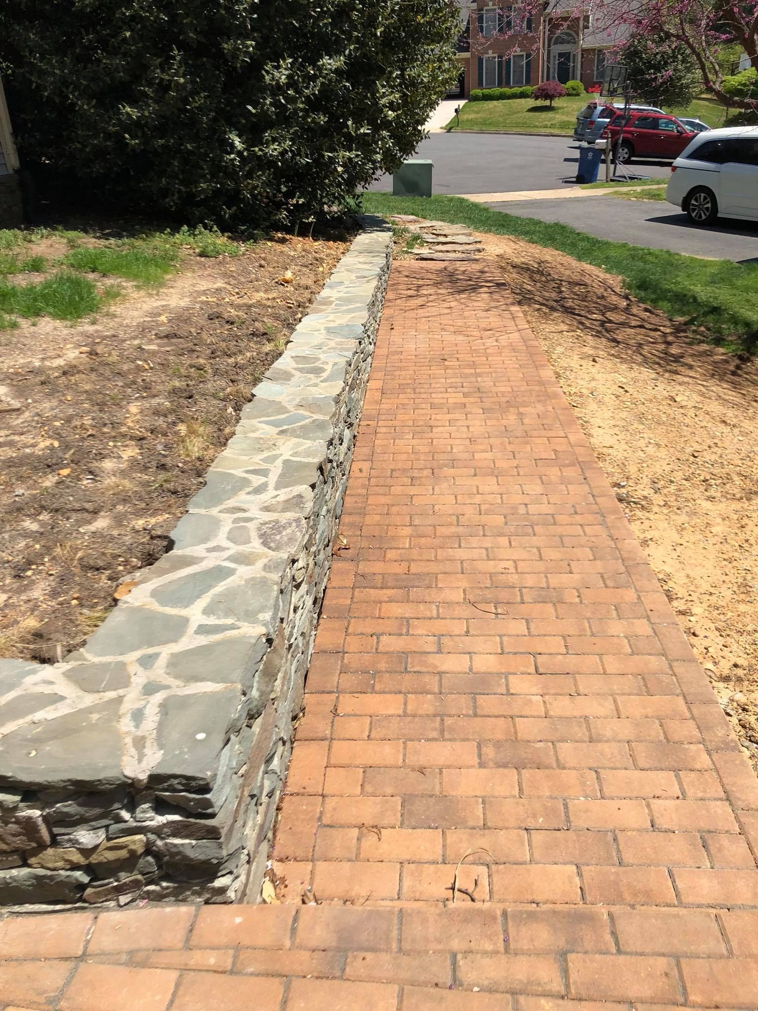 Brick pathway and stone wall next to grass. A person is standing near parked cars in the background.