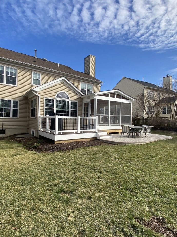 Backyard with a deck, screened porch, and stone patio. Beige house, blue sky, and green grass.