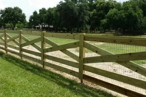 Wooden fence with X-shaped accents and wire mesh in a grassy field, trees in the background.