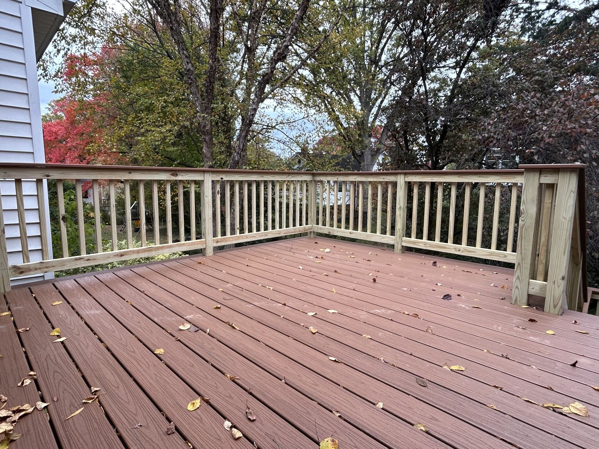 Wooden deck with railings, fallen leaves, and trees with autumn foliage.