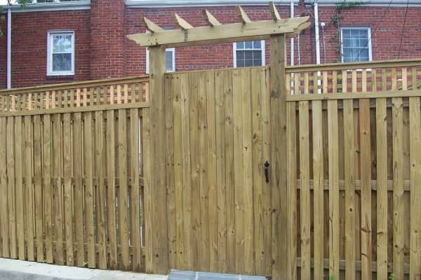Wooden fence and gate with pergola over the gate, against a brick building.