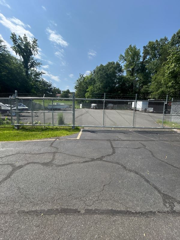 Chain-link fence enclosing a paved area with a closed gate; trees and sky in the background.