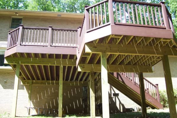 Multi-level wooden deck with brown railings, supported by posts. Exterior of a house with trees in the background.