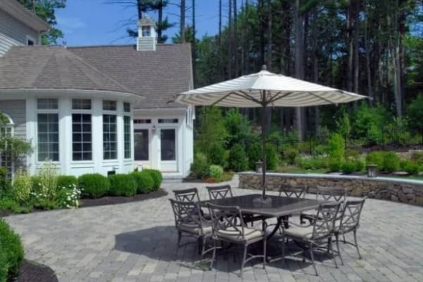 Patio with table, chairs, and umbrella outside a house. Green bushes and trees surround the area.