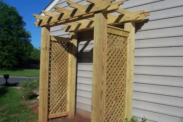 Wooden garden arbor with lattice side panels and a trellised top, next to a house.