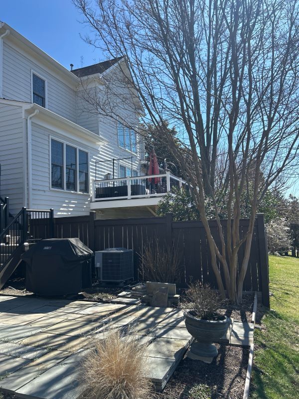 Backyard with house and deck. Features include a grill, air conditioner, and tree. Sunny, daytime setting.