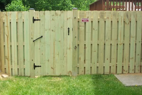 Wooden fence with gate, light green planks. Gate has black hinges and latch. Green grass and trees in background.