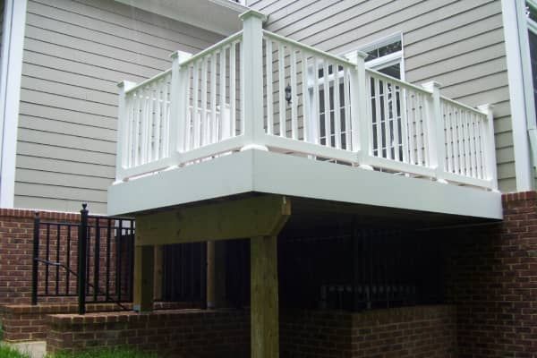 White deck with railing attached to a light gray house. Deck is above a brick foundation.