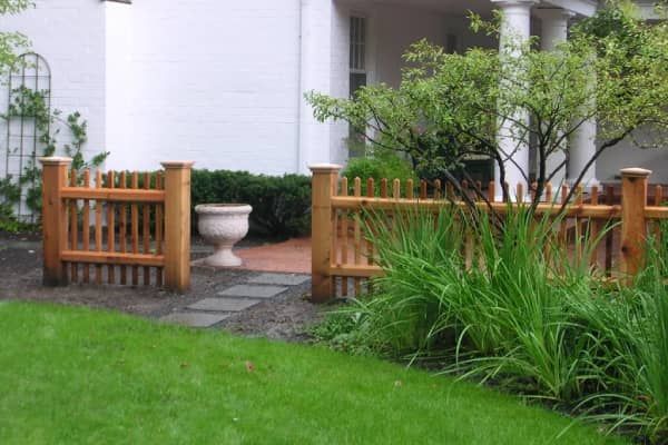 Wooden picket fence with large posts, in front of a white house with a green lawn.