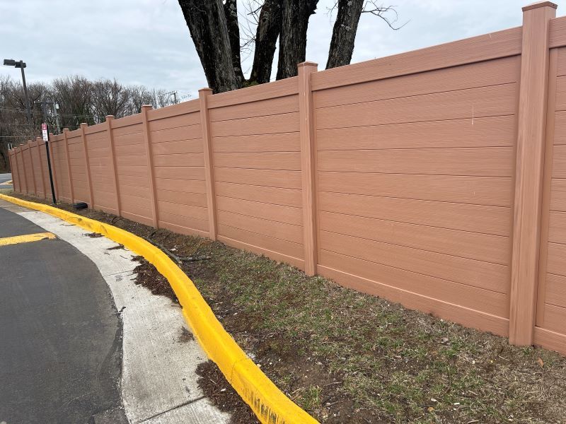 Brown privacy fence next to a yellow curb and grassy area. Trees and a cloudy sky are in the background.