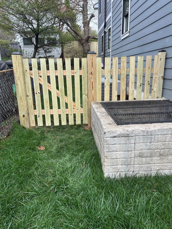 A wooden gate and fence section next to a stone planter box and a house with blue siding.
