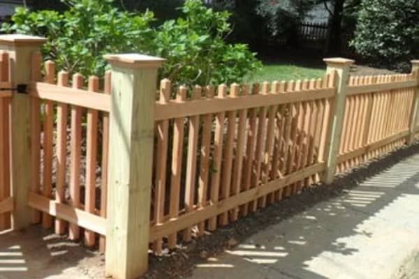 Wooden picket fence with gate, light brown color, in front of greenery.