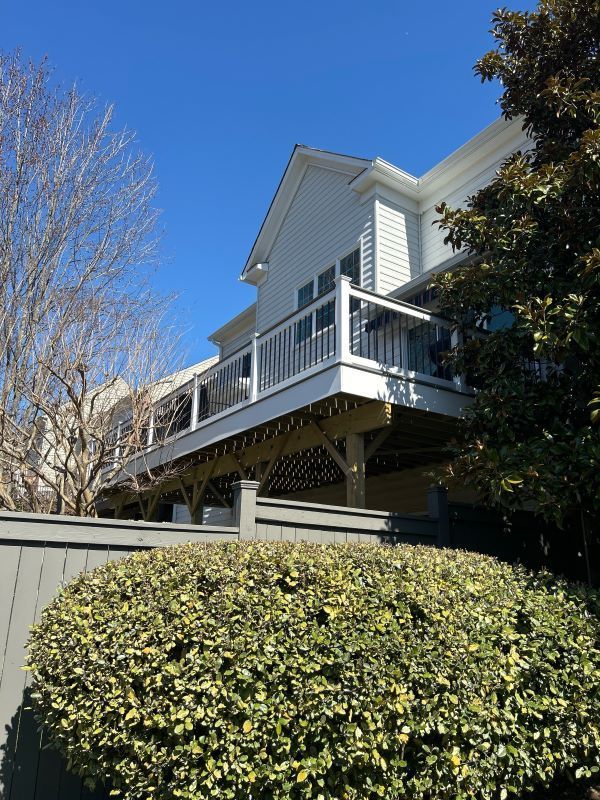 Elevated deck attached to a light blue house, with a fence and foliage in the foreground under a blue sky.