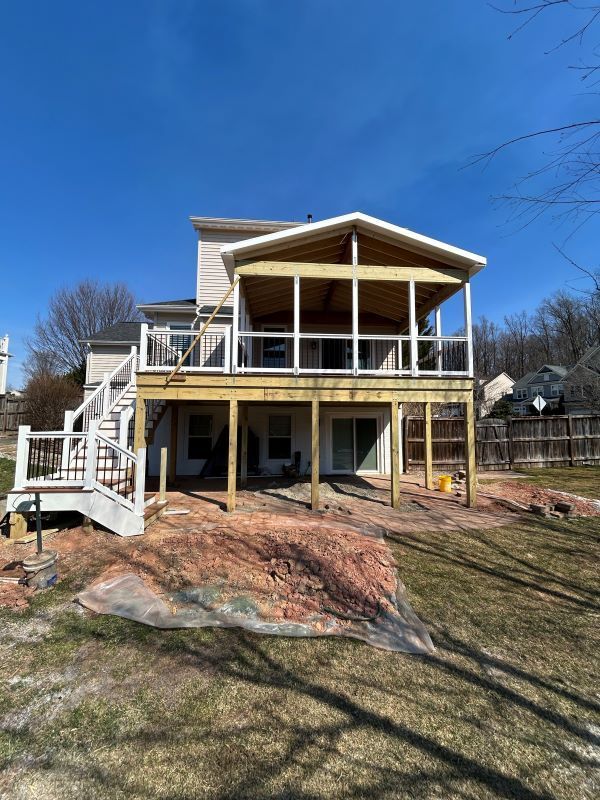 Two-story house with a wooden deck and screened porch. White stairs lead to the deck. Green grass and blue sky.