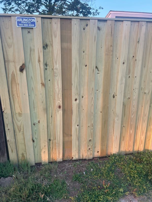 Wooden fence, vertical planks, light green with darker center plank, grass at base, blue company sign on top left.