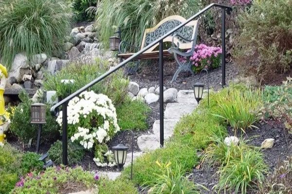A garden with stone steps, black handrail, bench, flowers, and solar lights.