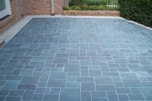 Gray stone patio with rectangular and square pavers. Brick wall and green bushes in the background.