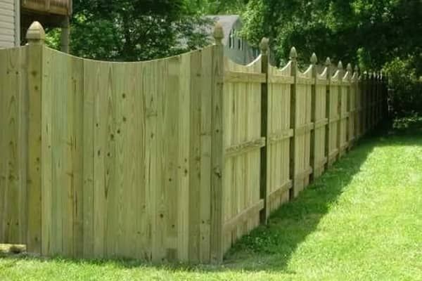Wooden fence in a backyard, with a curved top and decorative finials, bordering green grass.