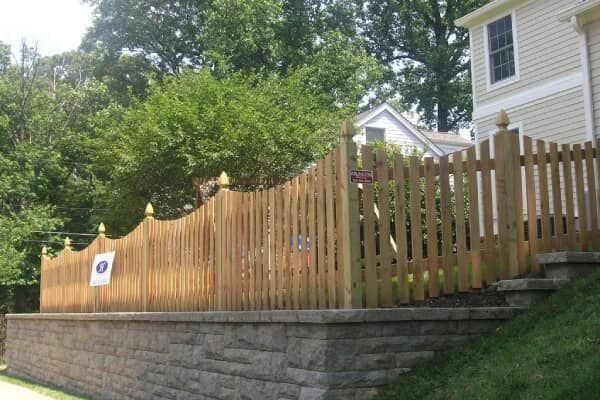 Wooden picket fence atop a retaining wall in front of a house.