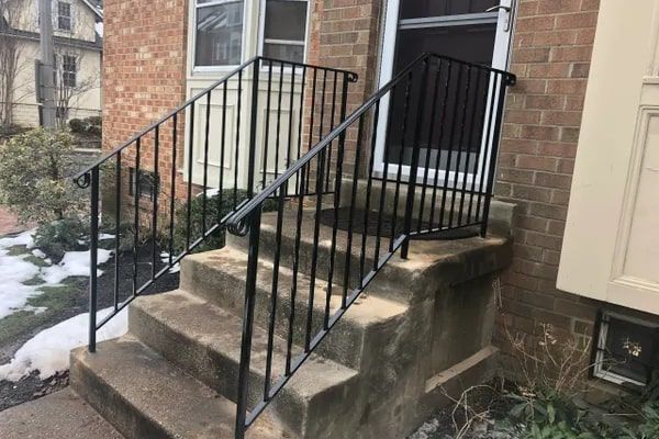 Black metal handrails on concrete steps leading to a brick building's entrance. Snow visible.