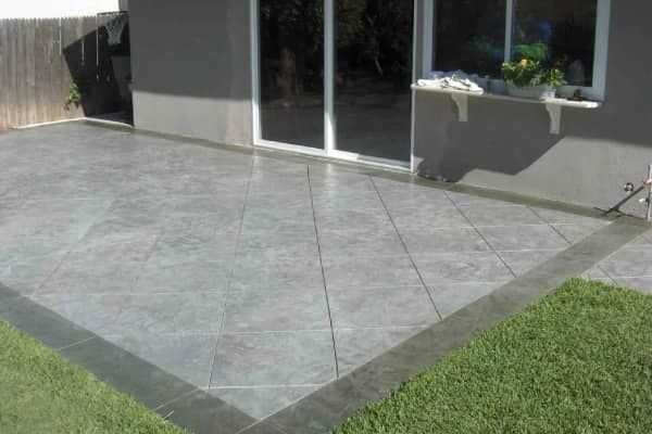 Grey patio with tile pattern and dark border next to a grassy lawn and a house with sliding glass doors.