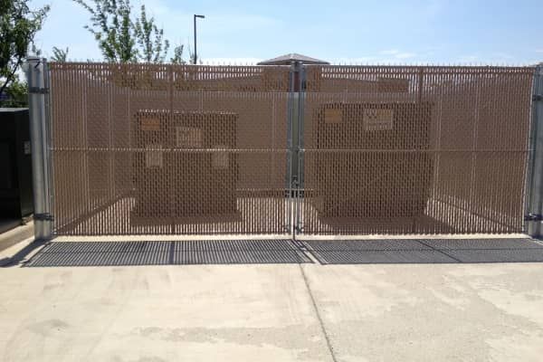Brown fenced enclosure with electrical equipment, secured by a gate, on a concrete surface.