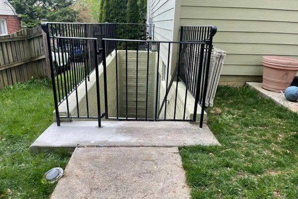 Black metal gate and railings enclosing basement window well on concrete pad.