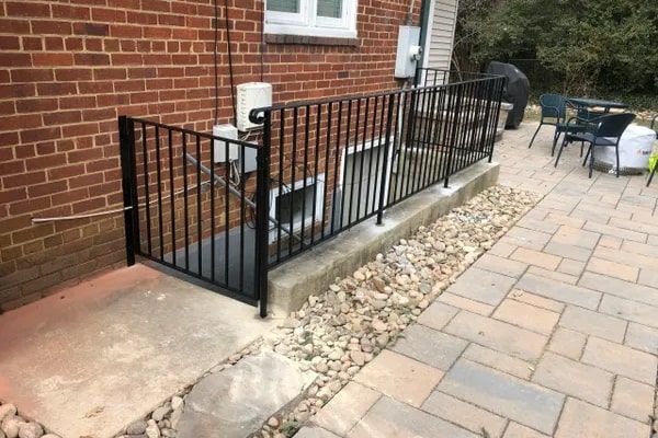 Black metal railing encloses basement window and walkway next to a brick building.