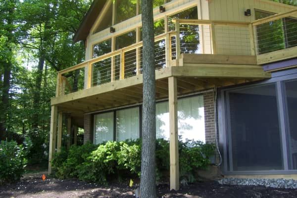 Wooden deck attached to a house with large windows, surrounded by trees and shrubbery.