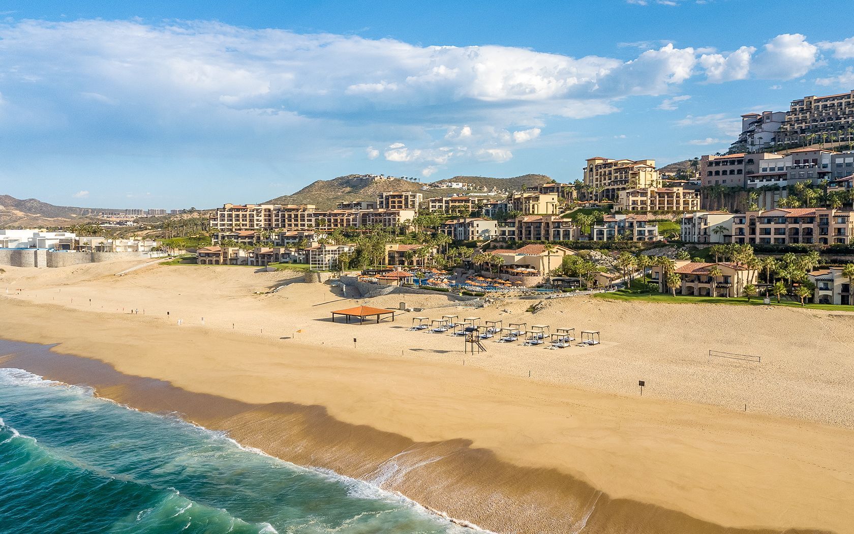 Sunny beach with turquoise waves, wide sand, umbrellas, and hillside resort buildings under a blue sky