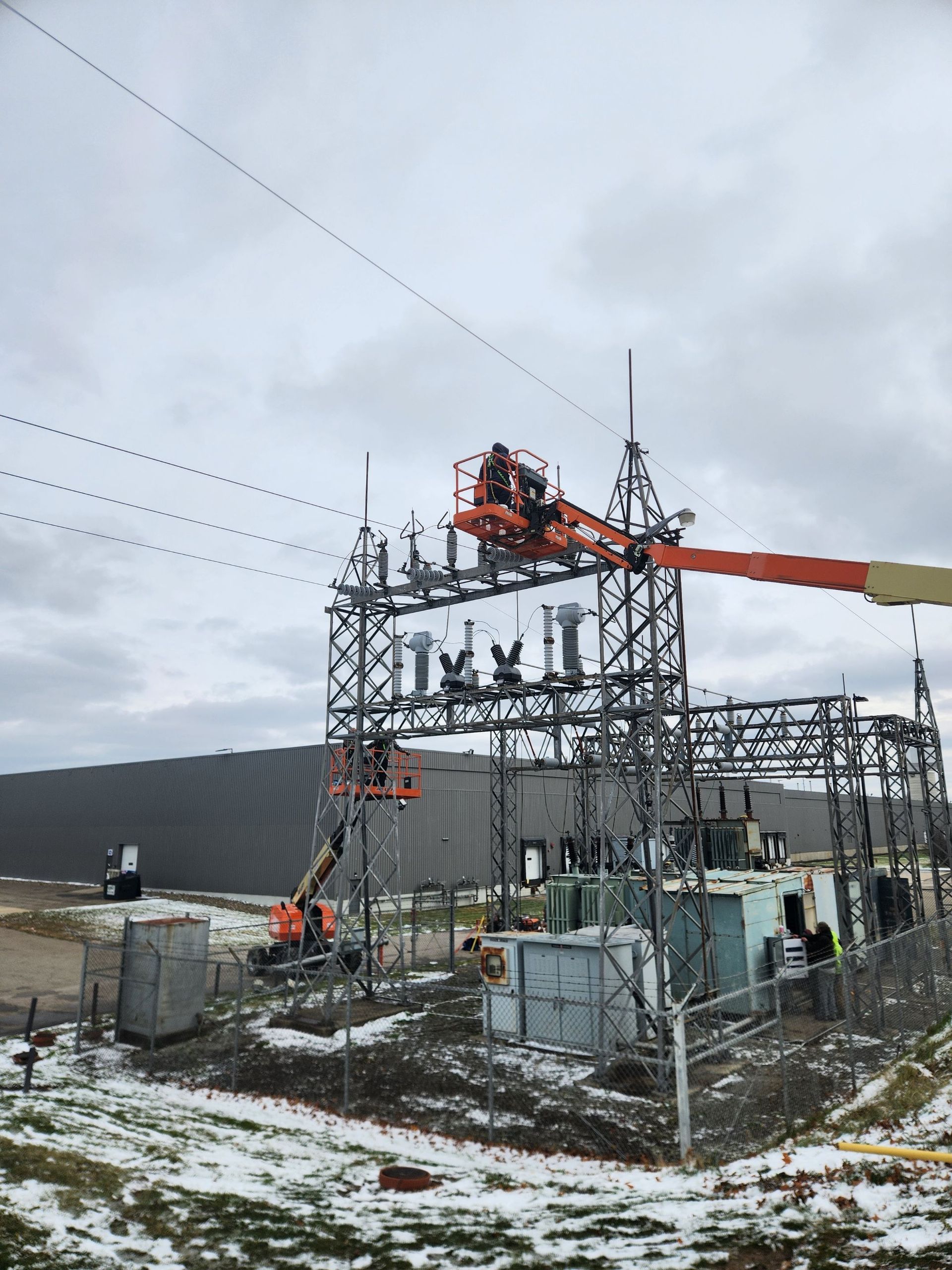 Workers in orange lifts at an electrical substation repairing power lines under an overcast sky.