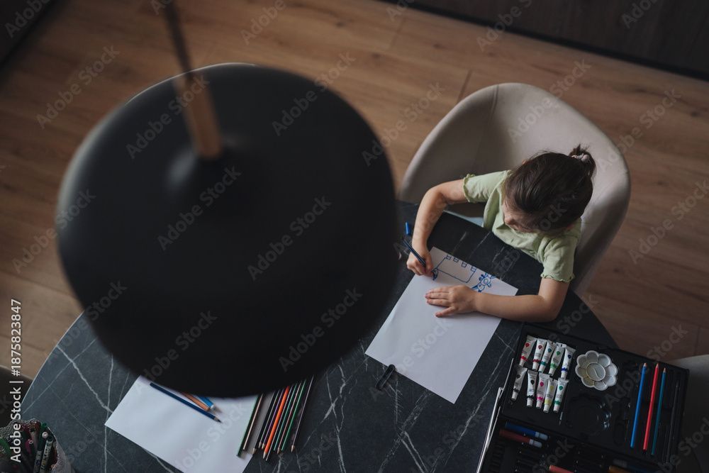 Child seated at table, drawing with colored pencils. Lamp hangs overhead.