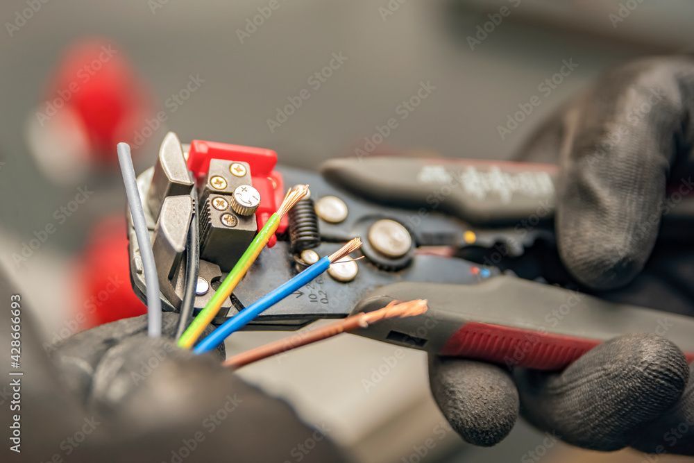 Gloved hands using wire strippers on electrical wires; blue, green, and brown exposed.