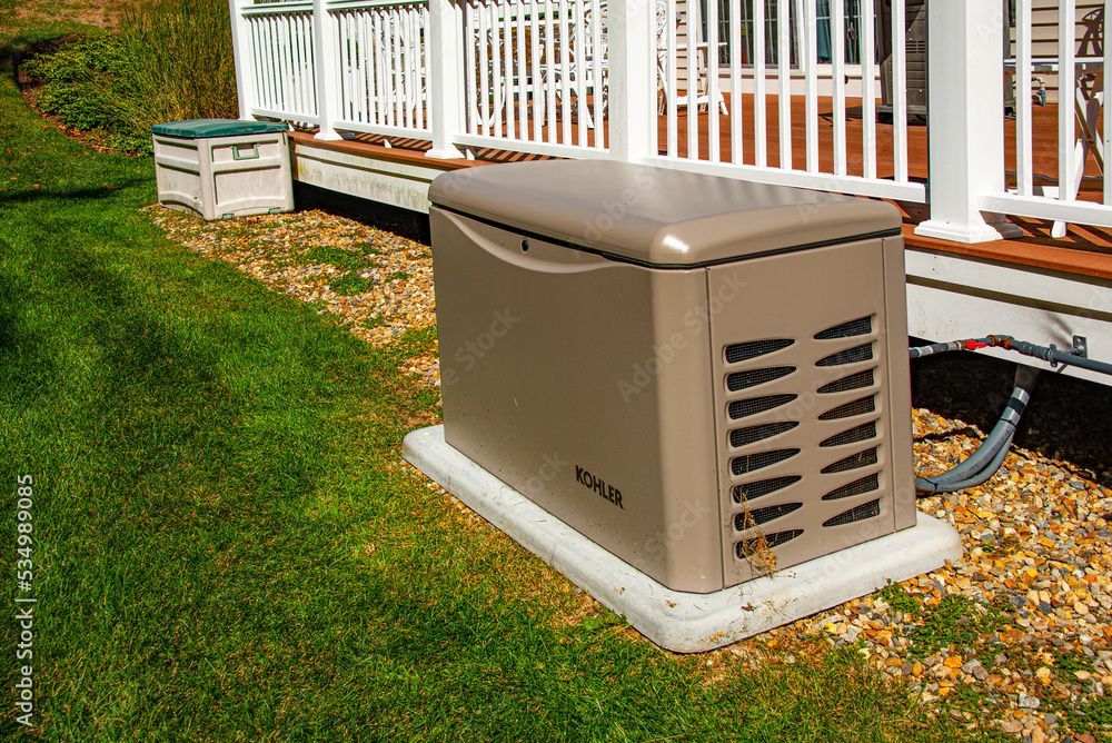 Tan home generator on concrete base beside a house with white railing and green lawn.