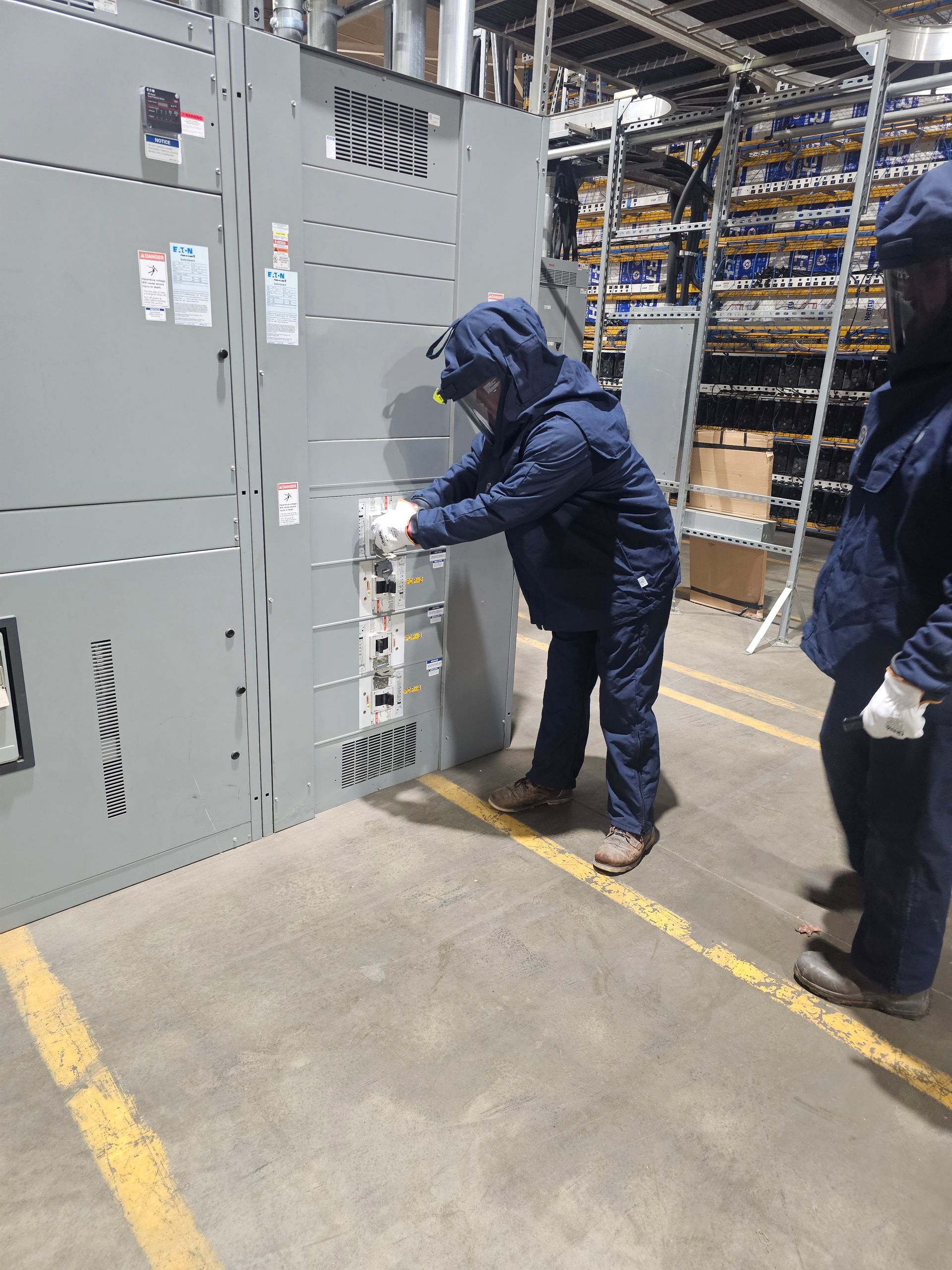 Person in work clothes operating electrical panel in a warehouse. Second person watches nearby.