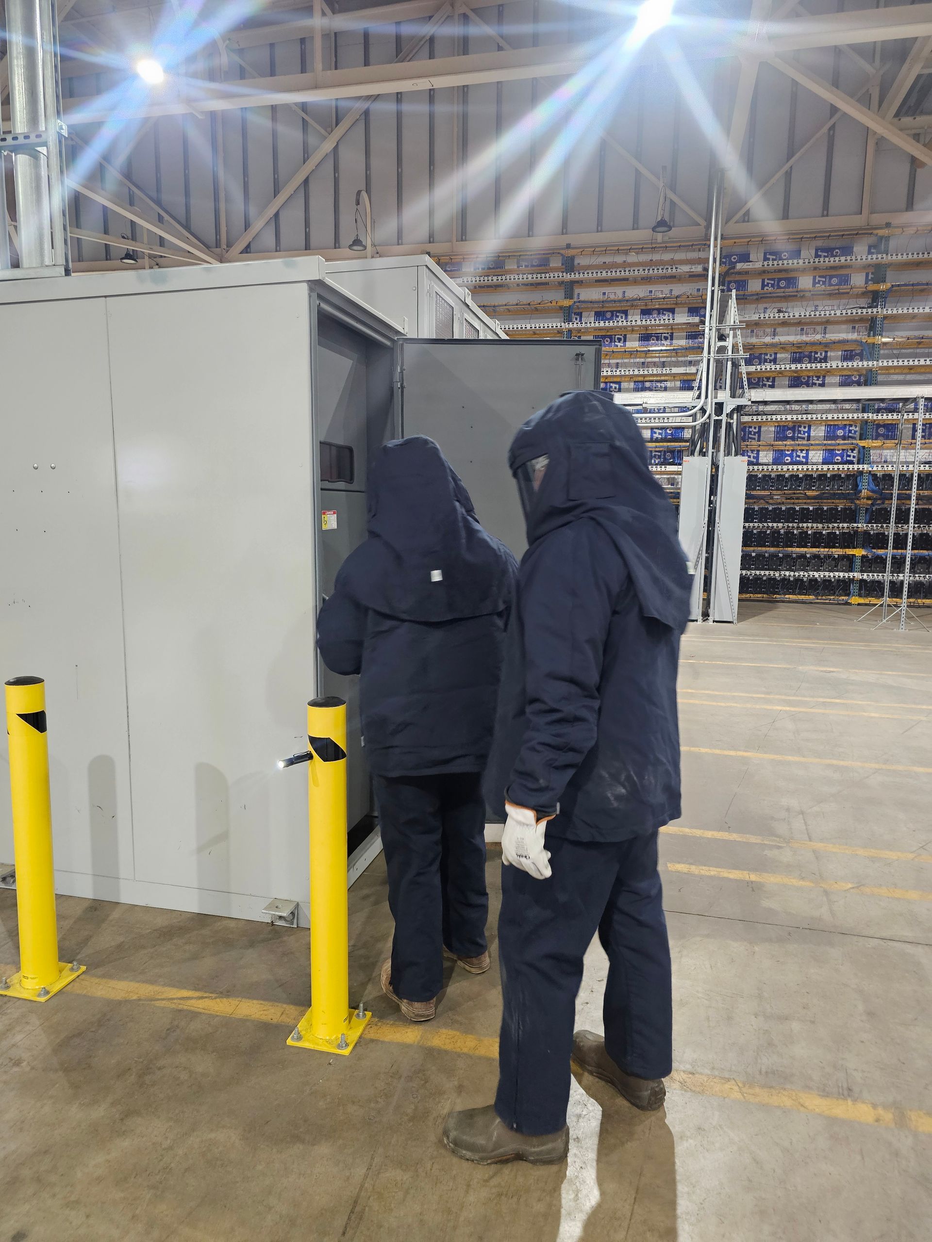 Two people in dark blue coveralls open an electrical cabinet, indoors, by two yellow bollards.