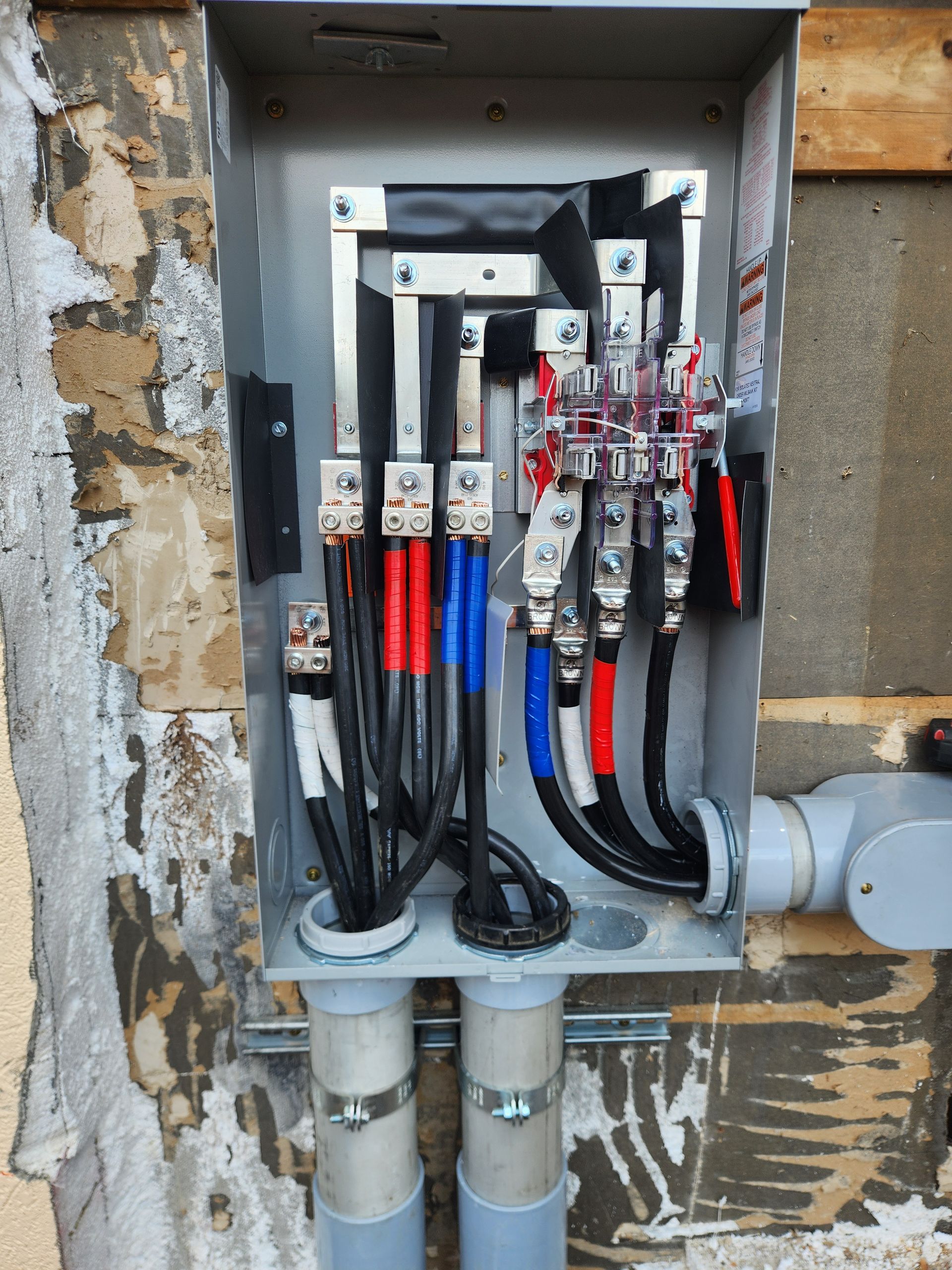Electrical panel with wires: black, red, blue, and white; housed in a metal box on a rough wall, pipes below.