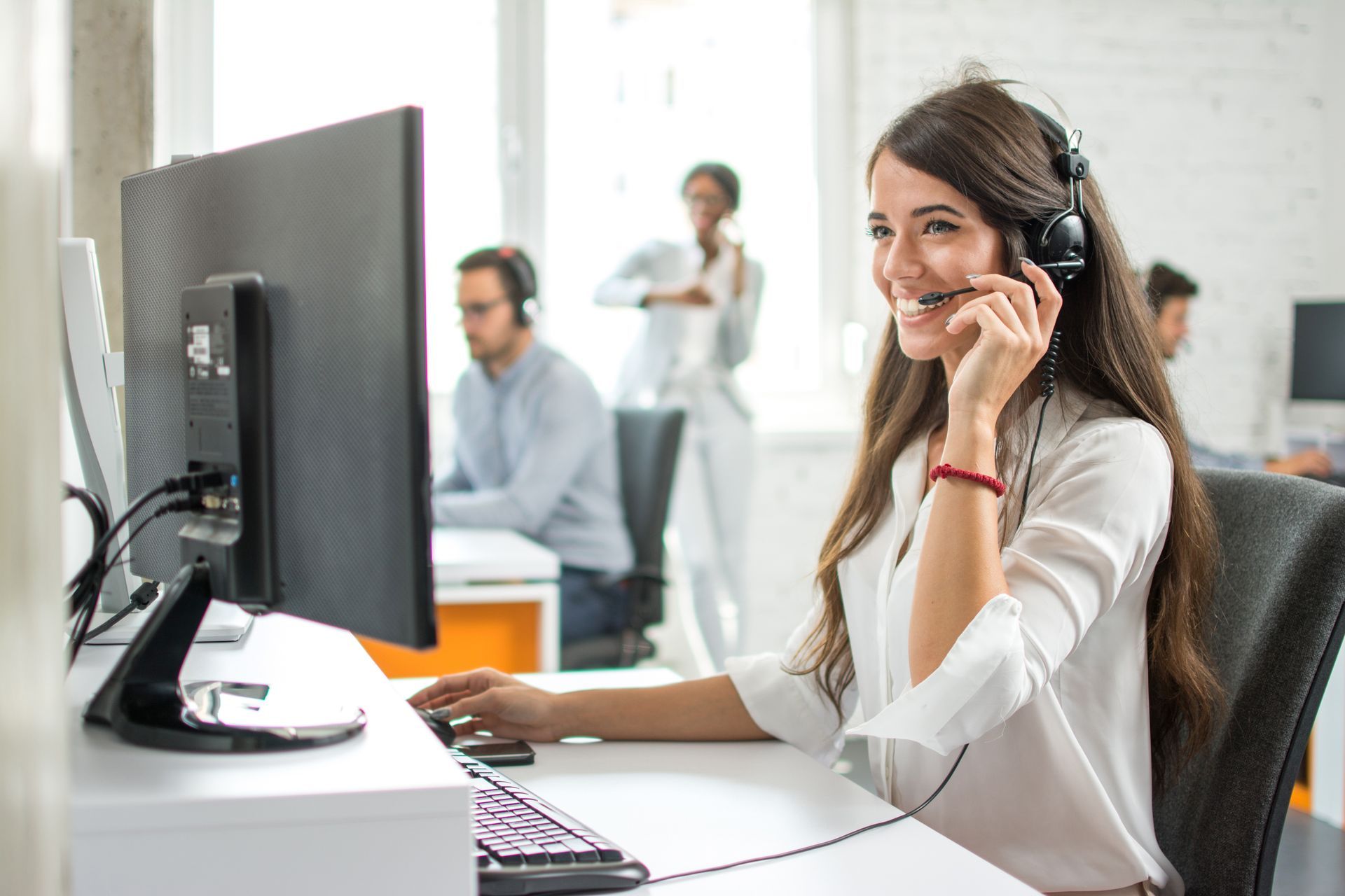 Woman with headset smiling, working at computer in an office, other workers in background.