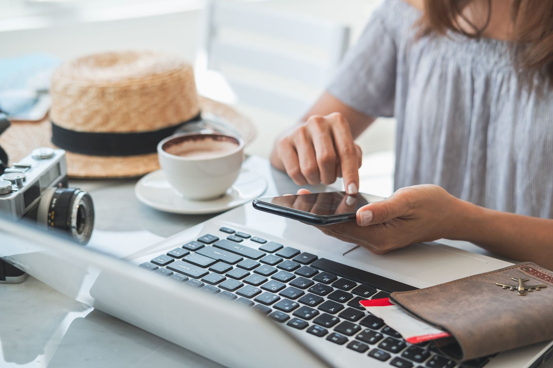 Woman using smartphone at a laptop with travel items: camera, hat, passport, and coffee cup.