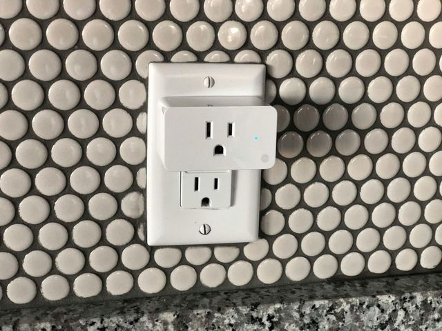 A white electrical wall outlet with a plug-in adapter attached, set against a background of white penny tiles.