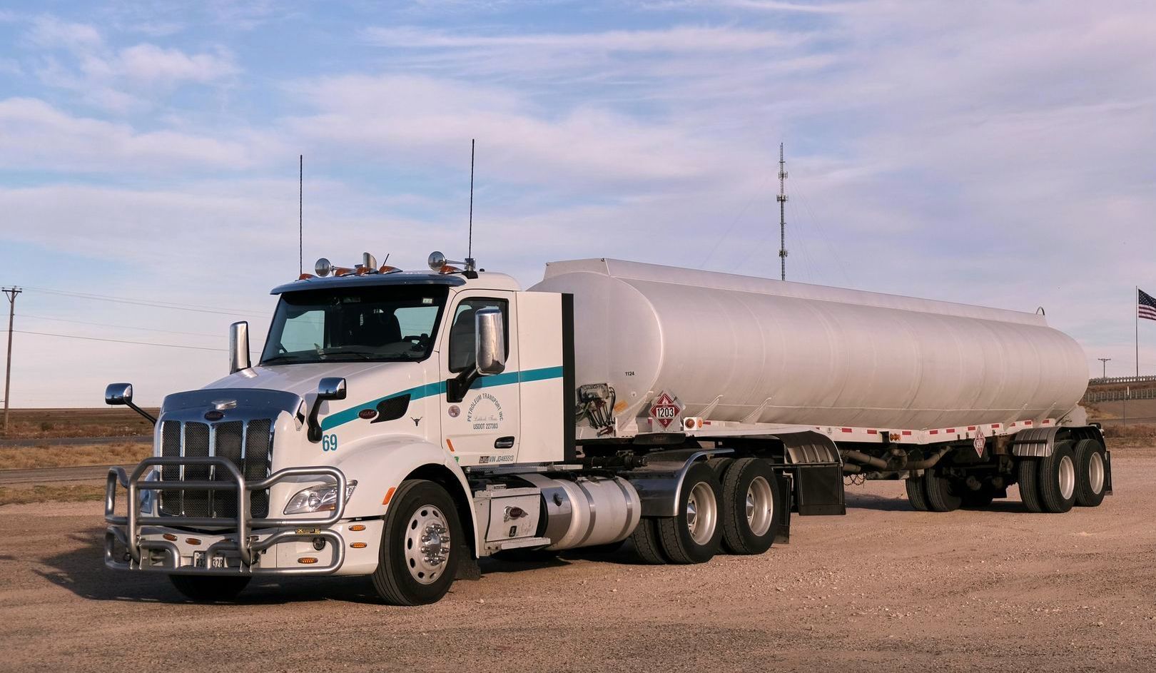 A white semi truck with a large tank on the back is parked in a gravel lot.