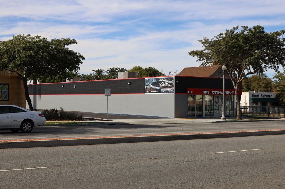 Exterior view of a commercial building with gray and red accents. Cars parked on street, blue sky.
