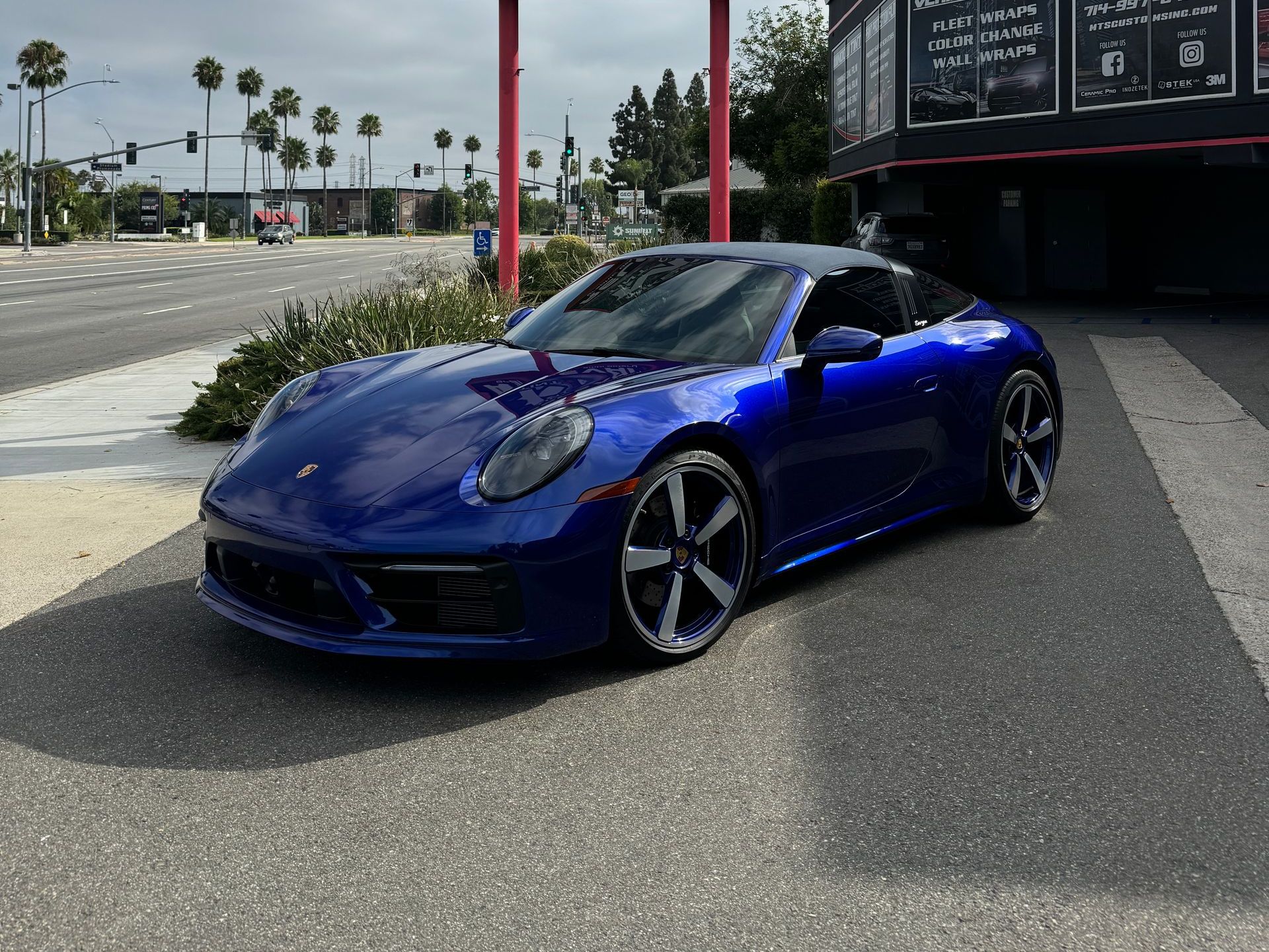 Blue Porsche 911 parked on a paved street. Black wheels, dark roof, sunny outdoor setting.