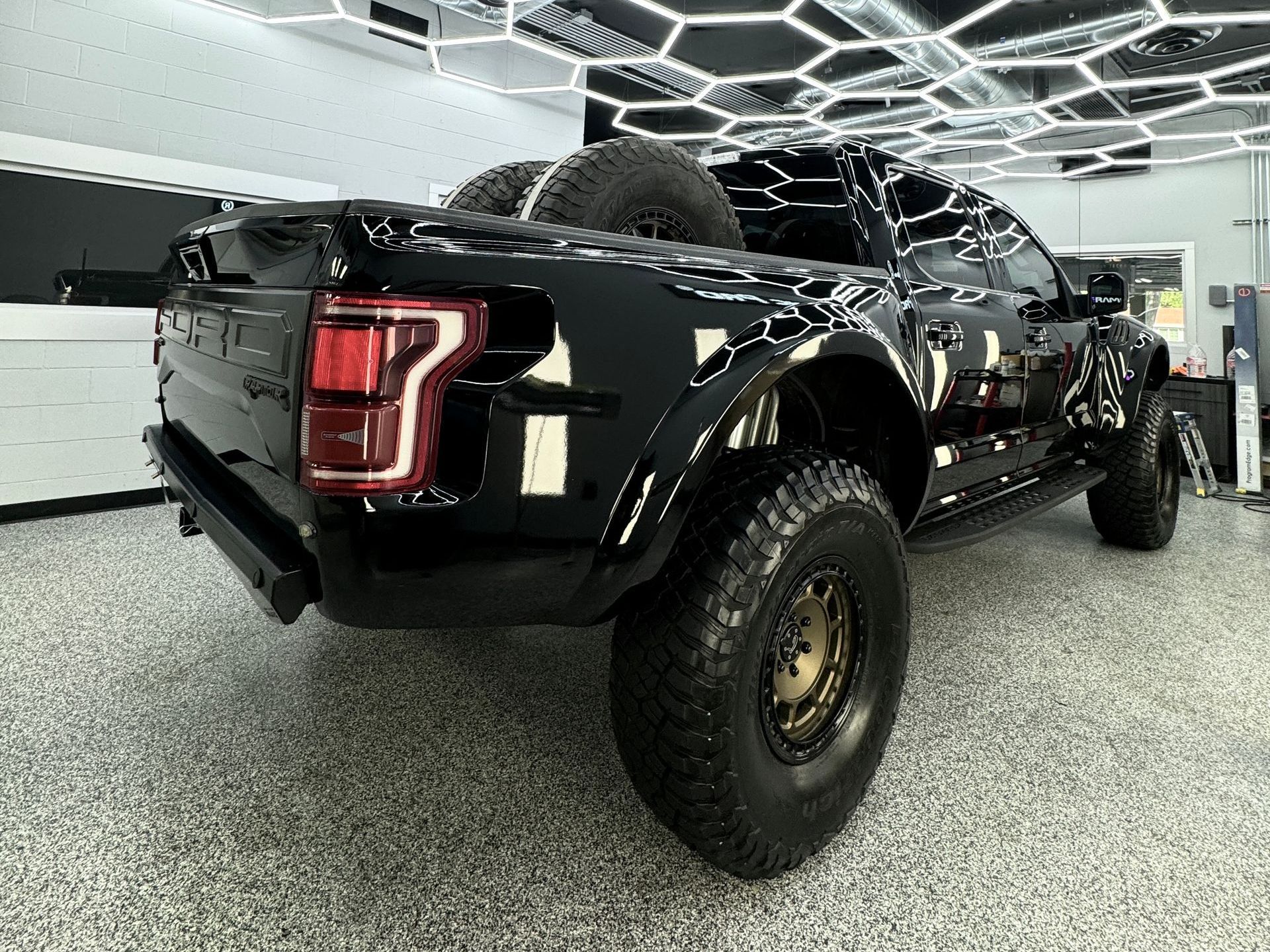 Black Ford Raptor truck with large tires, spare tires in bed, under a patterned ceiling.