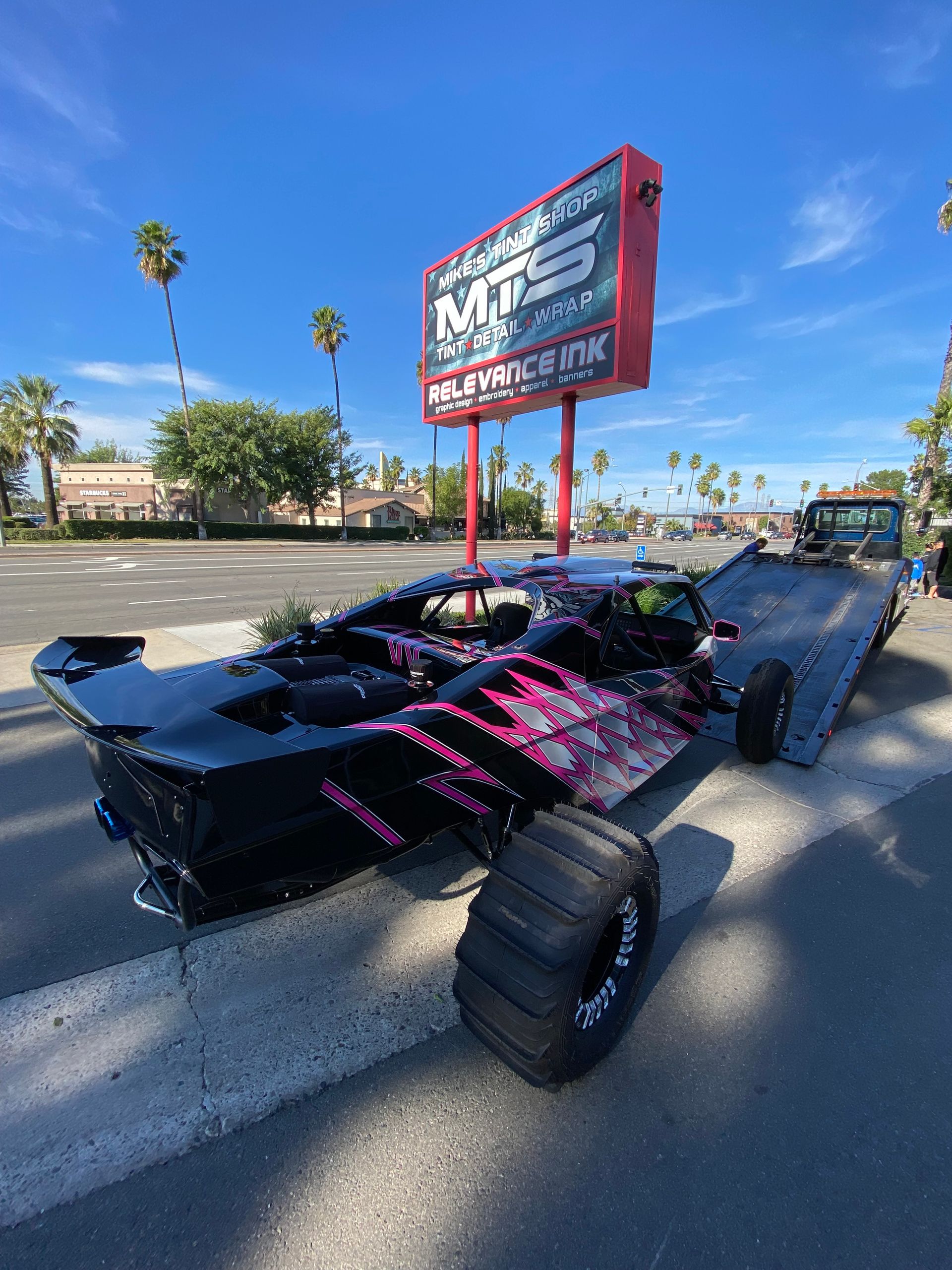 Black and pink custom drag car on asphalt with a sign in the background.