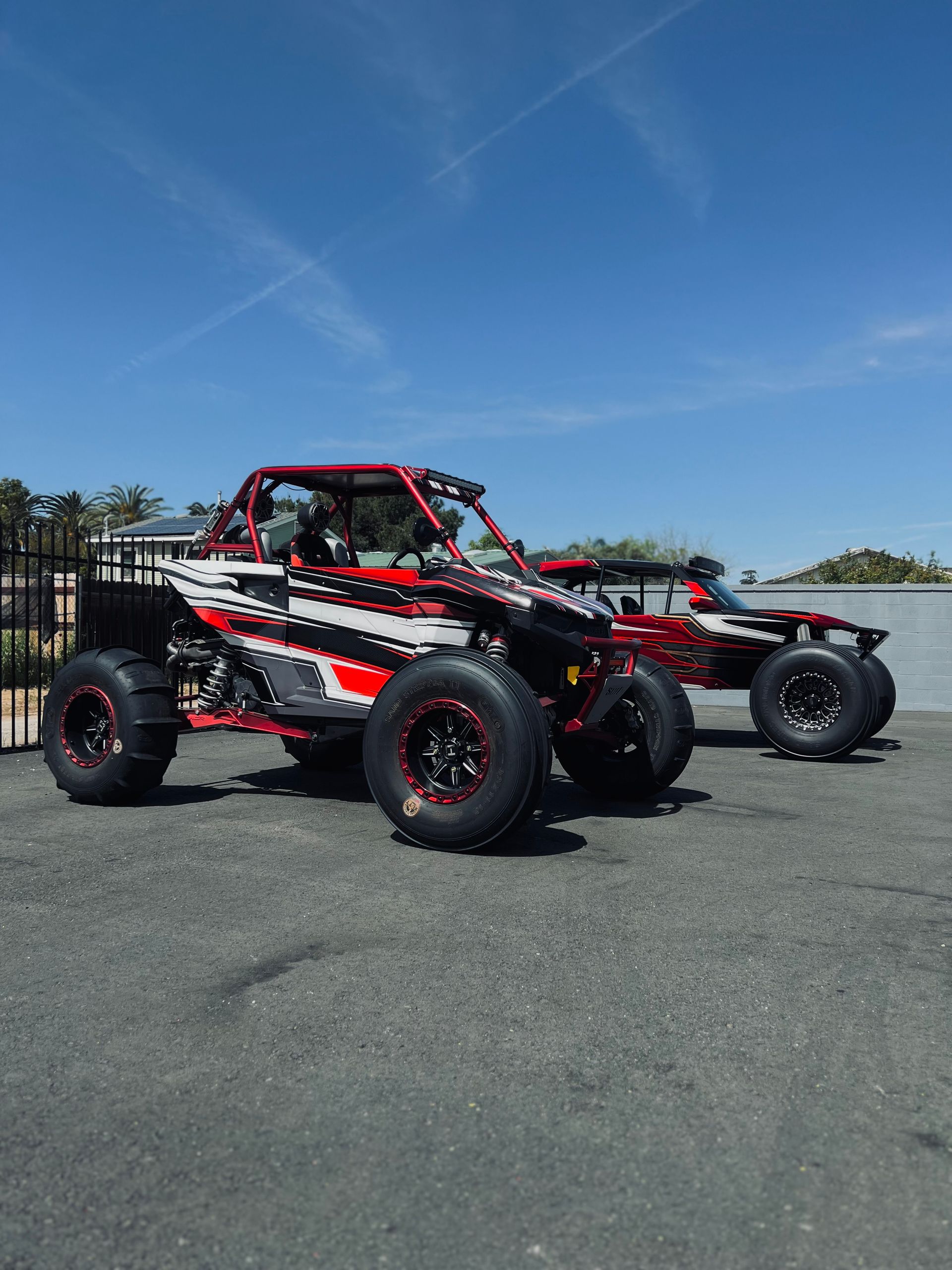 Two red, black, and white off-road vehicles parked on pavement, with a blue sky in the background.