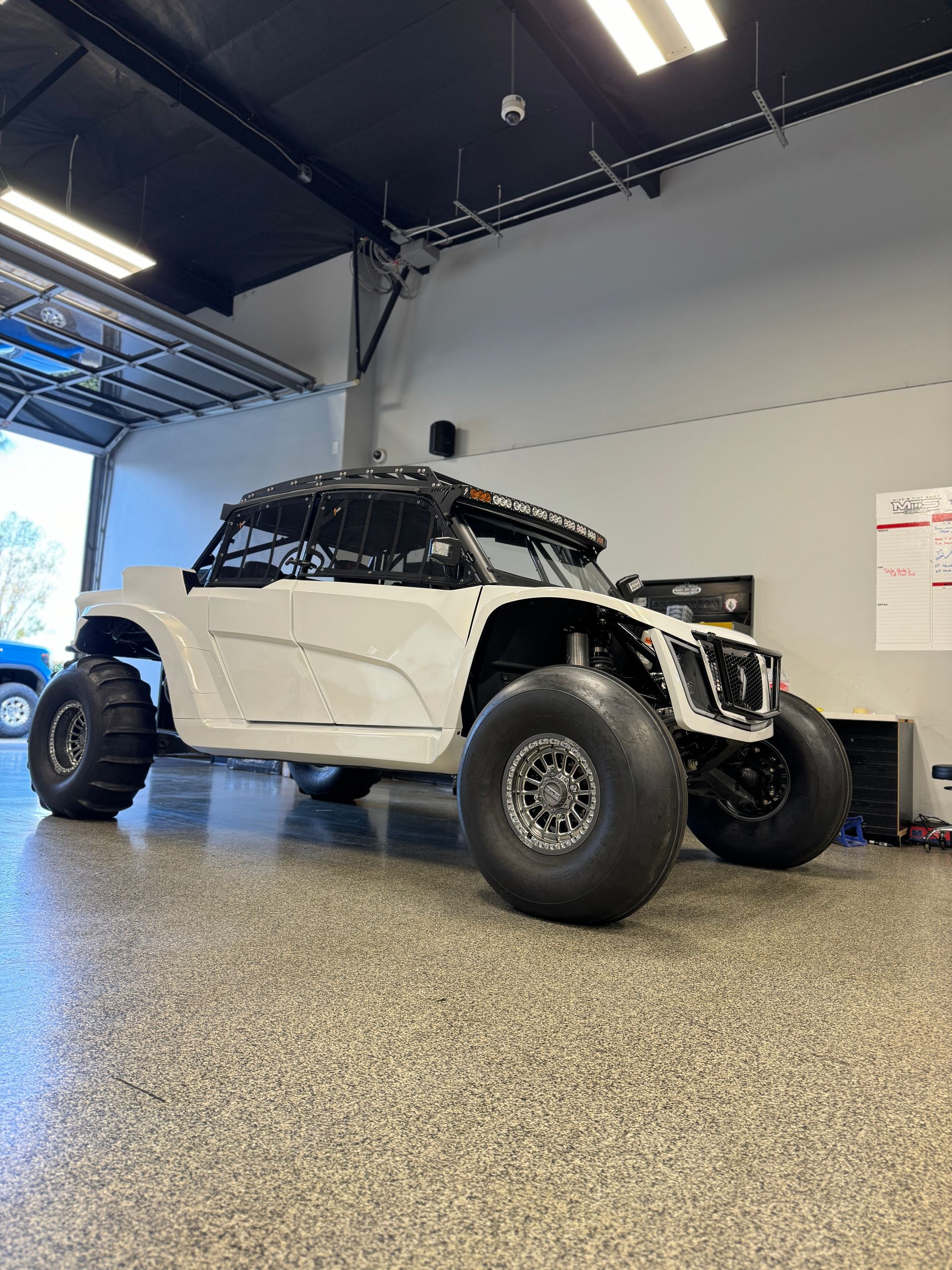 White off-road vehicle with large tires inside a garage with an open garage door.