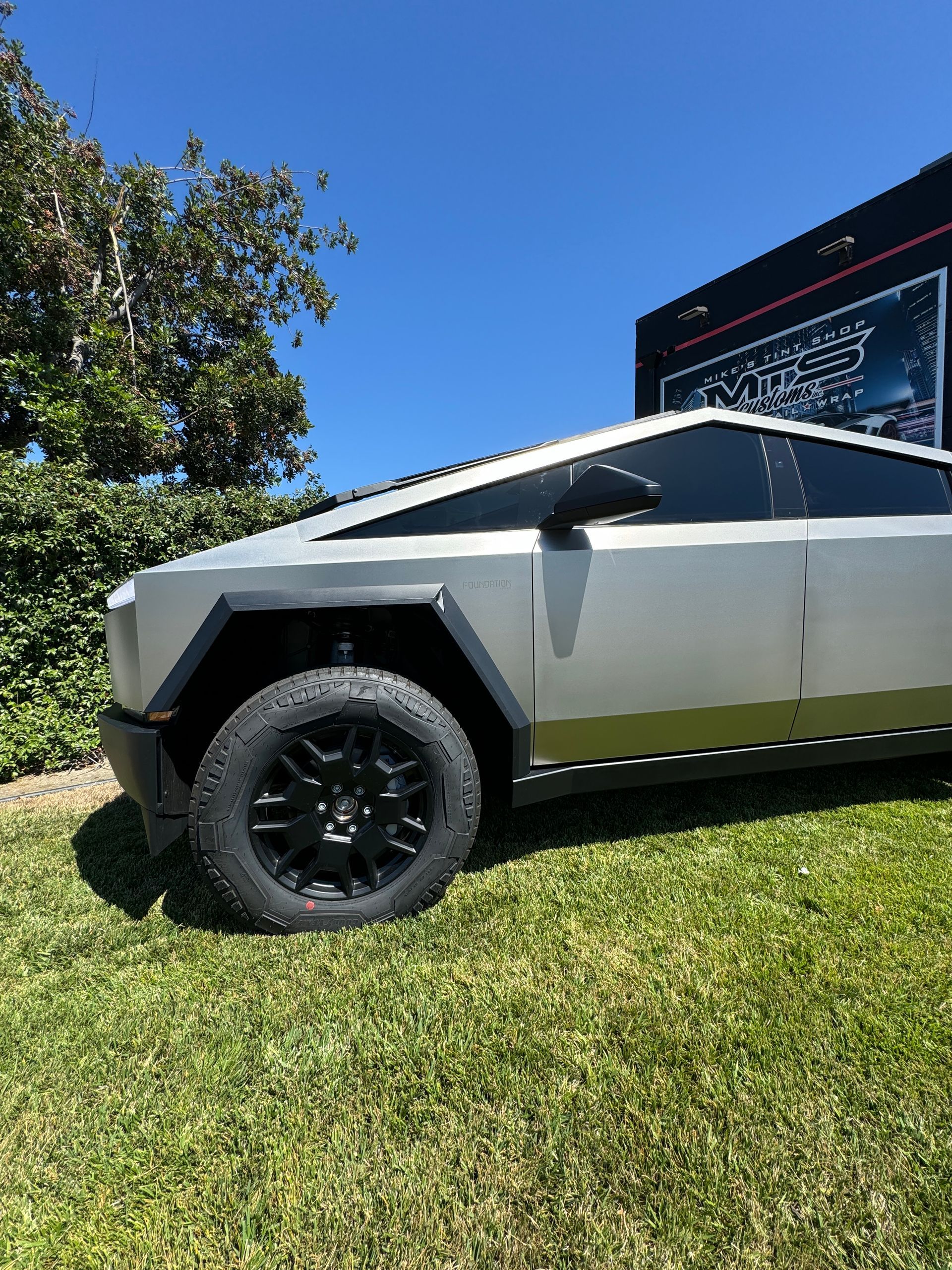 A silver tesla cybertruck is parked in the grass in front of a building.