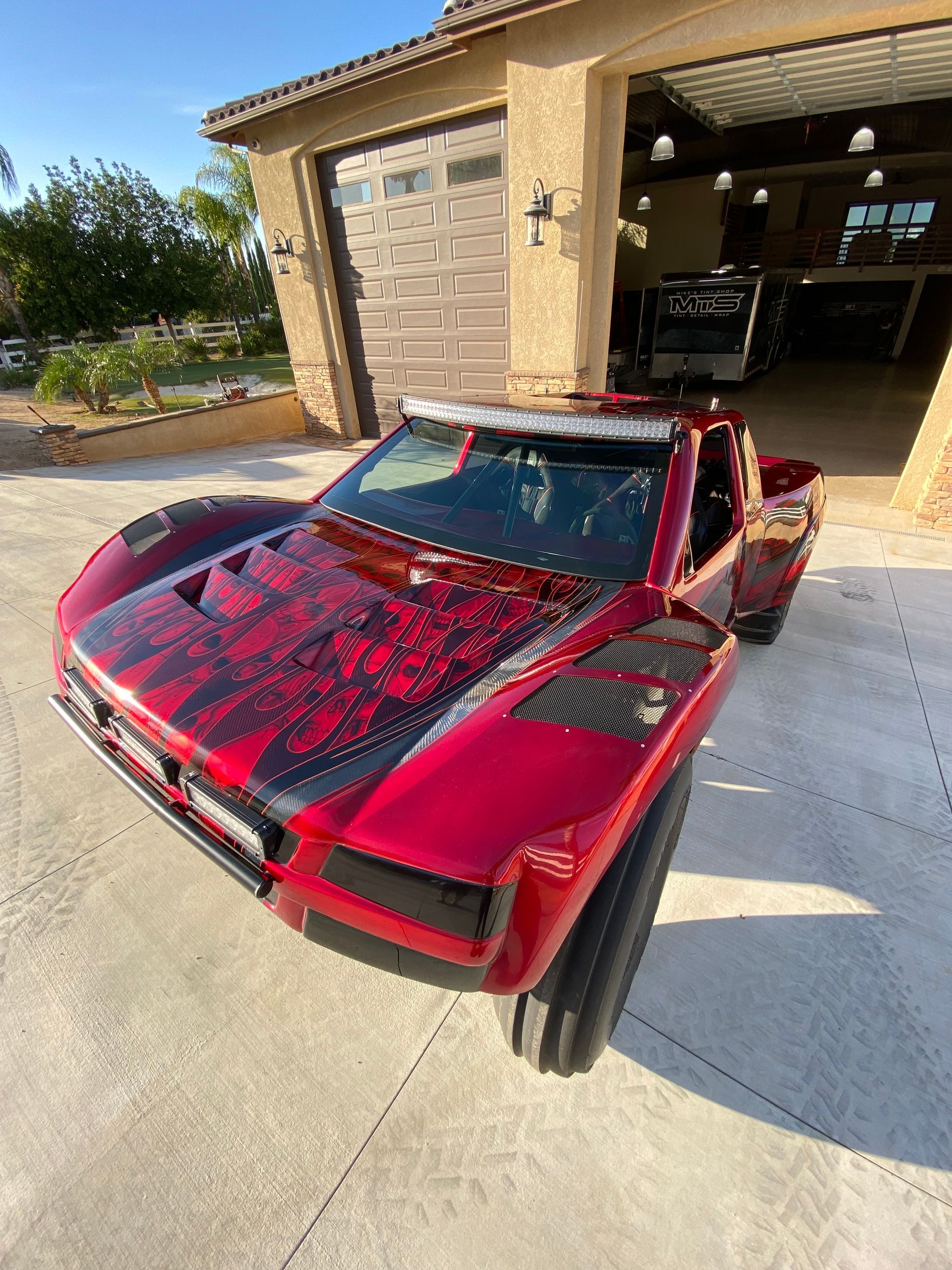 Red off-road truck with black accents parked in front of a garage, light bars on front.