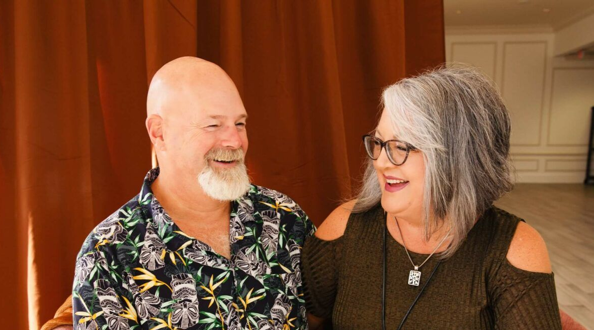 Smiling couple, man with bald head and floral shirt, woman with gray hair and glasses, sitting indoors.